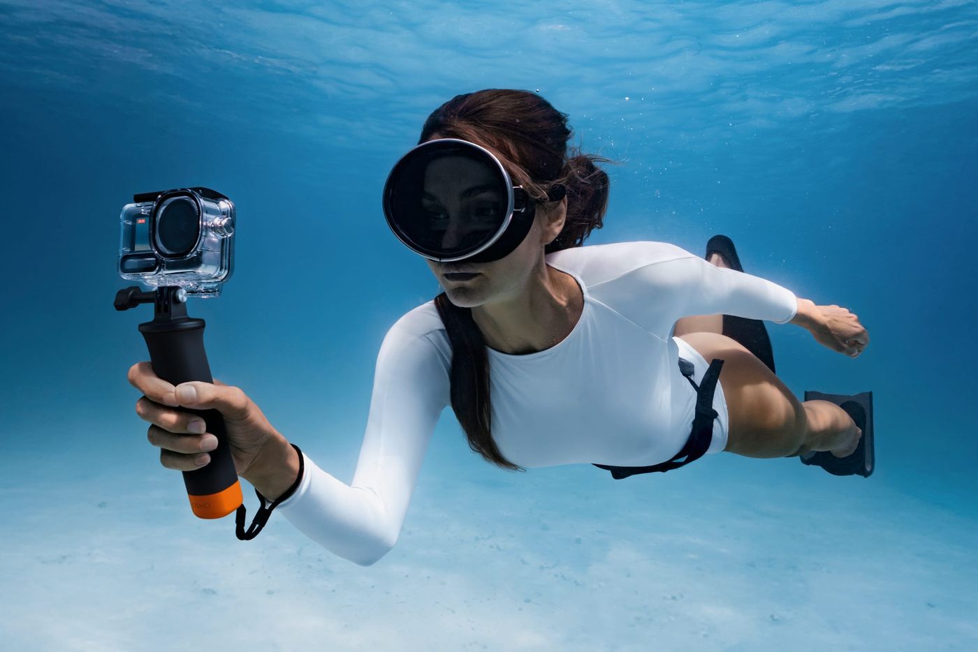 A snorkeller swimming over sand with a DJI Osmo Action camera in its waterproof case on a floating grip