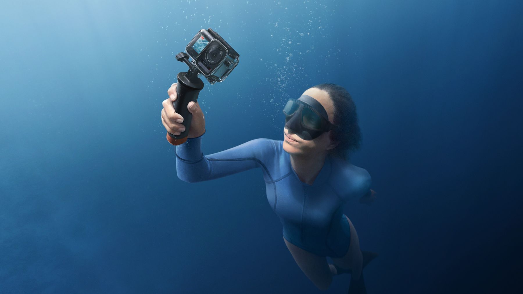 A freediver holding a DJI Osmo Action camera in its waterproof case on a floating grip, deep underwater